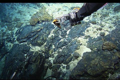 The manipulator arm of the Hercules ROV sampling rocks in the vicinity of the Atlantis Fracture Zone. Atlantic Ocean, Mid-Atlantic Ridge via NOAA