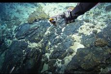 The manipulator arm of the Hercules ROV sampling rocks in the vicinity of the Atlantis Fracture Zone. Atlantic Ocean, Mid-Atlantic Ridge via NOAA